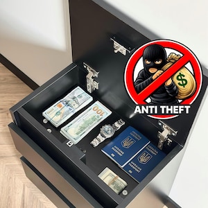 Pode incluir: Uma gaveta de armário preta aberta, revelando dinheiro, um relógio de pulso e dois passaportes ucranianos. A gaveta está rotulada como "ANTI THEFT" com um gráfico de um ladrão riscado. O armário é cinza escuro.