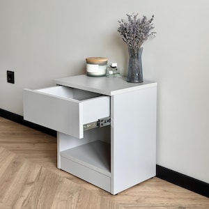 May include: A pale grey bedside table with an open drawer. The table has a lower shelf and is topped with a candle, perfume, and a vase of dried lavender. The drawer slides are visible. The unit is set against a neutral wall.