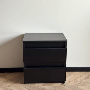 Pu&ograve; includere: Un comodino nero a tre cassetti o una piccola cassettiera. La superficie superiore &egrave; grigio scuro e i cassetti sono neri. L'unit&agrave; &egrave; rettangolare e poggia su un pavimento in legno.