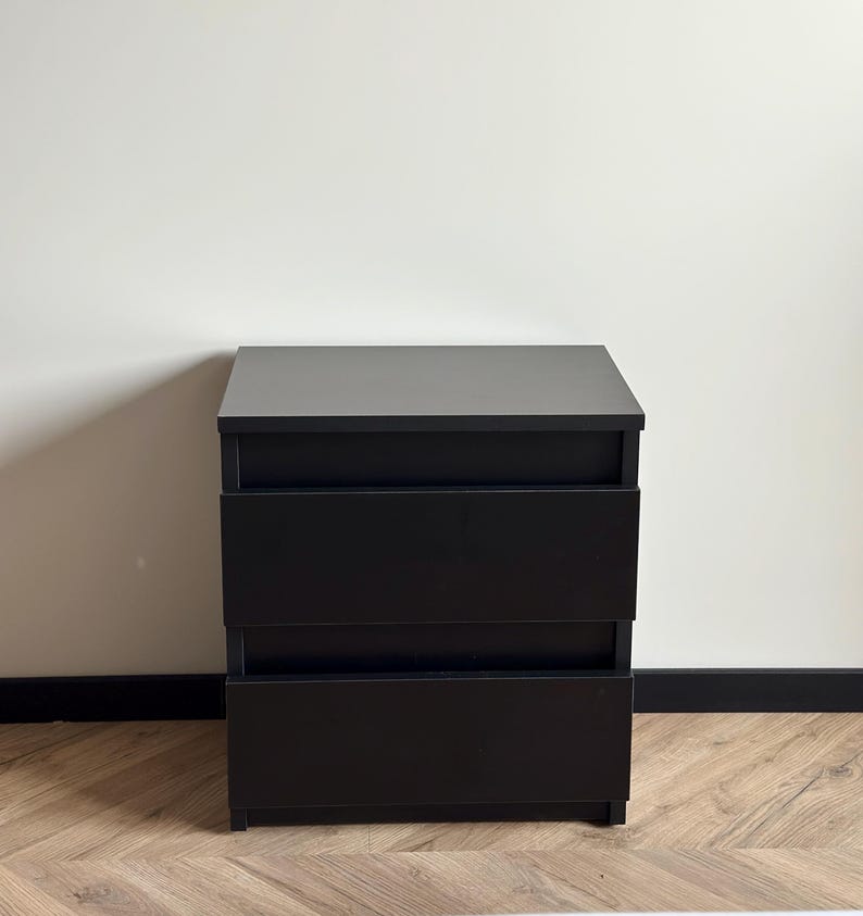 Pu&ograve; includere: Un comodino nero a tre cassetti o una piccola cassettiera. La superficie superiore &egrave; grigio scuro e i cassetti sono neri. L'unit&agrave; &egrave; rettangolare e poggia su un pavimento in legno.