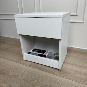 Puede incluir: Una mesita de noche blanca con una superficie superior lisa y plana y un cajón único situado sobre un compartimento de almacenamiento abierto. El estante abierto contiene una caja blanca rectangular, un objeto cilíndrico negro y varios billetes.