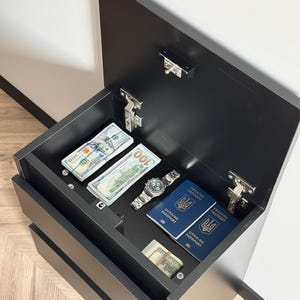 Pu&ograve; includere: Un mobile nero con la parte superiore aperta che rivela pile di banconote da cento dollari USA, un orologio da polso argentato e due passaporti ucraini blu. Il mobile ha due cassetti visibili ed &egrave; contro un muro bianco.
