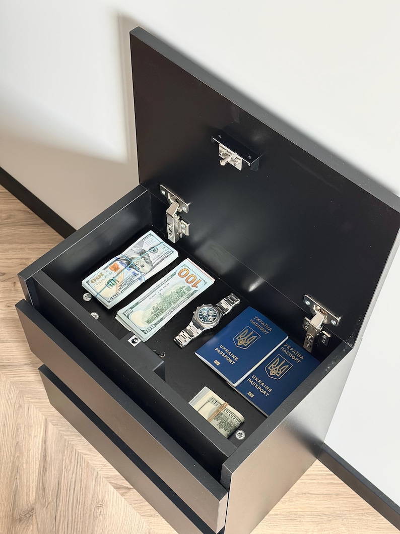 Pu&ograve; includere: Un mobile nero con la parte superiore aperta che rivela pile di banconote da cento dollari USA, un orologio da polso argentato e due passaporti ucraini blu. Il mobile ha due cassetti visibili ed &egrave; contro un muro bianco.