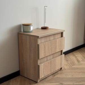 Pu&ograve; includere: Un comodino in legno marrone chiaro con due cassetti. Una candela in un barattolo di vetro con coperchio in legno e un porta incenso minimalista sono sopra. Il comodino &egrave; su un pavimento in legno.