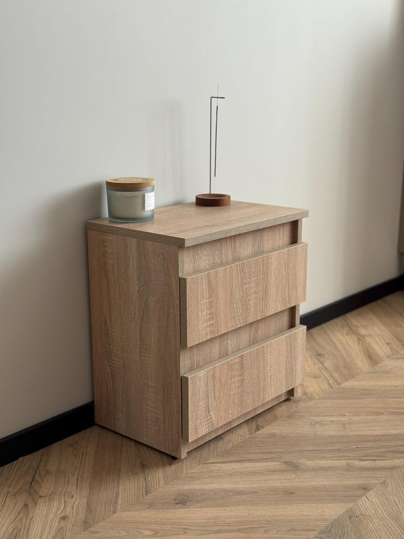 Pu&ograve; includere: Un comodino in legno marrone chiaro con due cassetti. Una candela in un barattolo di vetro con coperchio in legno e un porta incenso minimalista sono sopra. Il comodino &egrave; su un pavimento in legno.