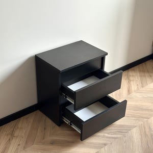 Pu&ograve; includere: Un comodino nero con due cassetti, uno parzialmente aperto. I cassetti hanno interni bianchi. &Egrave; appoggiato a una parete chiara e a un pavimento in legno. Design semplice e moderno.