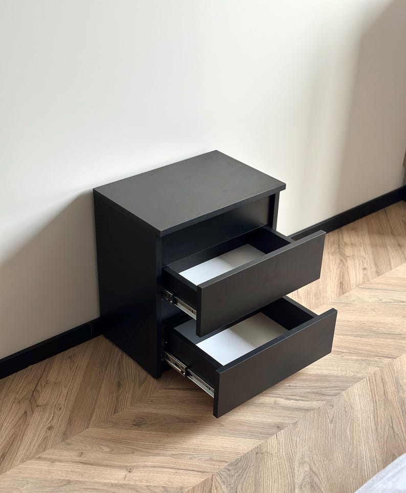 Pu&ograve; includere: Un comodino nero con due cassetti, uno parzialmente aperto. I cassetti hanno interni bianchi. &Egrave; appoggiato a una parete chiara e a un pavimento in legno. Design semplice e moderno.