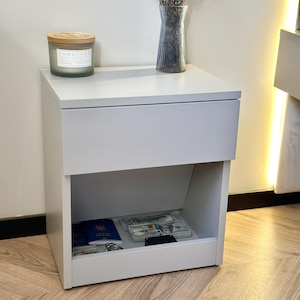 May include: A light grey bedside table with a single top drawer and an open lower storage compartment. The top surface holds a green candle with a wooden lid and a clear ribbed glass vase containing dried lavender flowers.