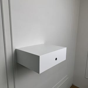 May include: A white, floating wall-mounted nightstand or shelf with a single drawer. The drawer has a small, round, black handle. The shelf is mounted on a white wall.