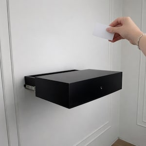 May include: A black floating drawer mounted on a white wall. The drawer is rectangular with a smooth, matte finish. A hand is holding a white card above the drawer. The drawer is open, revealing the drawer slides.