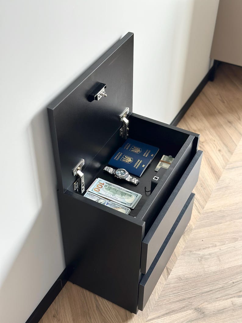 May include: A black, wall-mounted cabinet with an open top drawer revealing a passport, cash, and a wristwatch. The cabinet is mounted on a wall with a light wood floor visible.