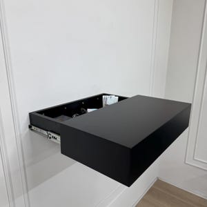 May include: A black floating shelf with a hidden drawer, mounted on a white wall. The drawer is partially open, revealing contents like keys and money. The shelf's design is minimalist and modern, offering concealed storage.