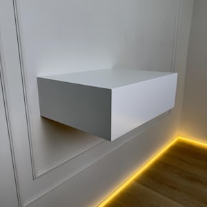 May include: A white floating shelf mounted on a white wall with a yellow LED light strip along the baseboard.