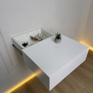 May include: A white floating nightstand with an open drawer revealing keys and cash. A small potted plant sits on the nightstand's surface. The nightstand is mounted on a white wall above a wooden floor.
