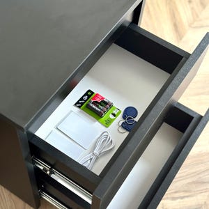 Pu&ograve; includere: Un mobile nero a due cassetti con cassetti aperti che rivelano vari oggetti. Il cassetto superiore contiene batterie, portachiavi e carte bianche. &Egrave; visibile anche un cavo bianco. Il mobile &egrave; su un pavimento in legno.
