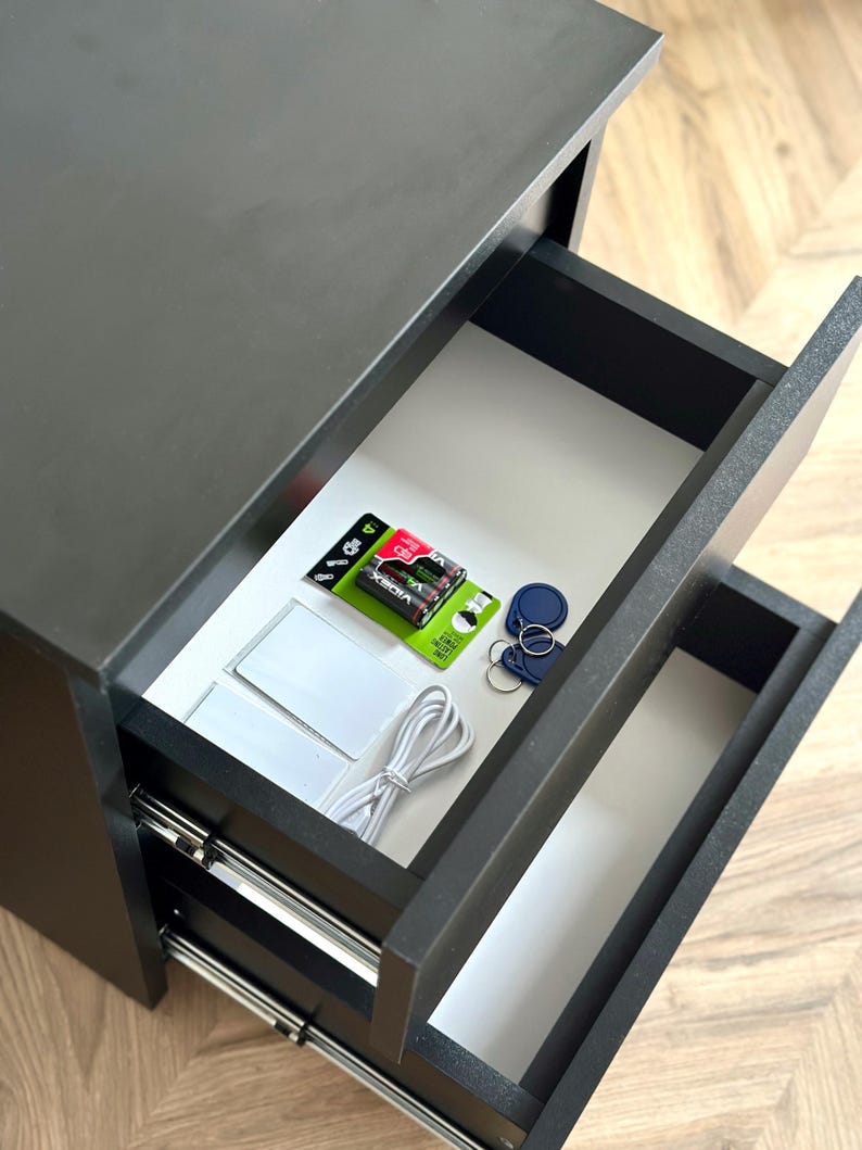 Pu&ograve; includere: Un mobile nero a due cassetti con cassetti aperti che rivelano vari oggetti. Il cassetto superiore contiene batterie, portachiavi e carte bianche. &Egrave; visibile anche un cavo bianco. Il mobile &egrave; su un pavimento in legno.