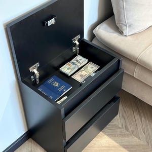 Könnte beinhalten: Ein schwarzer Nachttisch mit zwei Schubladen und einem oberen Fach. Die Oberseite ist geöffnet und zeigt Geldbündel, einen ukrainischen Reisepass und ein Schloss. Der Nachttisch steht neben einem beigen Sofa.