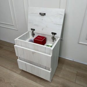 May include: A white wood-grain bedside table with two lower drawers and a top compartment that opens upwards on metal hinges. Inside the open top section sits a small red box labelled Guardo and two paper banknotes. The unit has a clean finish.