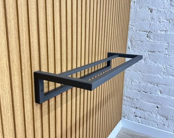 Könnte beinhalten: Ein mattschwarzes Metallregal, das an einer vertikalen Wand mit Holzpaneelen befestigt ist. Das Regal hat ein rechteckiges Design mit zwei parallelen Stangen. Die Wand ist hellbraun mit vertikalen Rillen. Im Hintergrund ist eine weiße Backsteinmauer zu sehen.