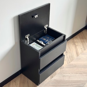 May include: A matte black bedside table with a hinged top compartment that opens to reveal a hidden storage space. Inside the compartment are a stack of one hundred dollar bills, a silver wristwatch, and a blue passport. Two drawers are below.