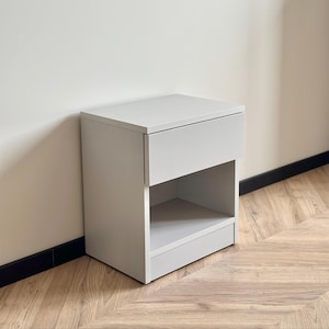 Puede incluir: Una mesita de noche gris claro con un cajón y un estante abierto. La mesita de noche es de madera y está sobre un suelo de madera. El diseño es moderno y minimalista, adecuado para un dormitorio. Medidas en centímetros.
