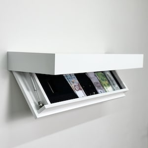 Puede incluir: Un estante flotante rectangular de color blanco montado en la pared con un compartimento de almacenamiento oculto. El panel frontal está inclinado, revelando un interior forrado en negro que contiene varios billetes y una bisagra metálica.