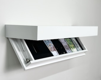 May include: A white rectangular wall-mounted floating shelf with a hidden storage compartment. The front panel is tilted open, revealing a black interior lining containing several paper banknotes and a small metallic support hinge on the left side.