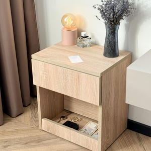 Pode incluir: Uma mesa de cabeceira em tom de madeira clara com uma gaveta e uma prateleira aberta. Uma pequena lâmpada rosa com uma lâmpada redonda, um frasco de perfume e um vaso de lavanda estão em cima. A prateleira contém um relógio, anéis, um telefone e dinheiro.
