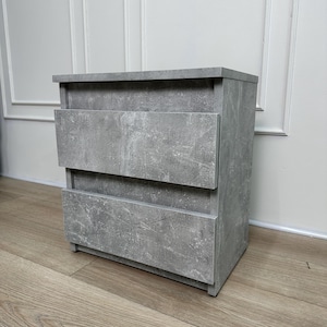 Pu&ograve; includere: Un comodino grigio a due cassetti con finitura materica effetto cemento. L'armadietto rettangolare ha un piano piatto e poggia su un pavimento in legno. I cassetti hanno un design semplice e moderno.