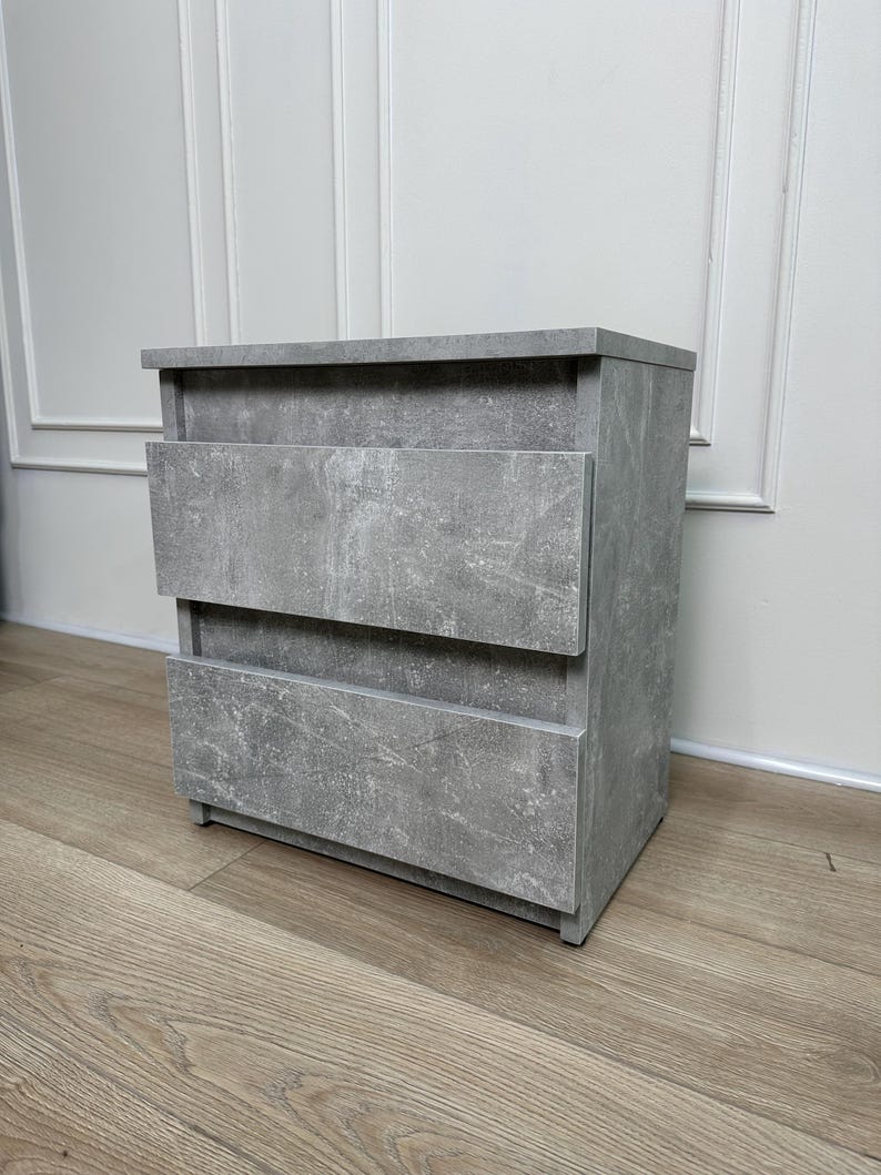 Pu&ograve; includere: Un comodino grigio a due cassetti con finitura materica effetto cemento. L'armadietto rettangolare ha un piano piatto e poggia su un pavimento in legno. I cassetti hanno un design semplice e moderno.