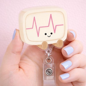 May include: A cute, cartoon-style badge reel featuring a smiling heart monitor design with a pink heartbeat line. The badge reel has a clear plastic clip and a retractable cord for easy use.