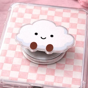 May include: A white cloud-shaped phone grip with a smiling face and brown feet. The phone accessory is attached to a clear phone case with a pink and white chequered pattern.