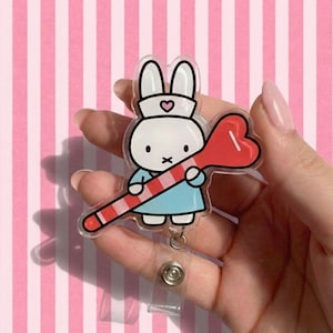 May include: A clear acrylic badge reel featuring a cartoon bunny wearing a nurse's hat and holding a red heart-shaped lollipop with a red and white striped stick. The bunny is wearing a blue dress. The badge reel has a clear plastic strap and a metal clip.
