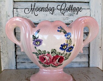 May include: A pink ceramic vase with two handles and a pedestal base. The vase is decorated with hand-painted flowers in shades of red, blue, and purple. The text "Moondog Cottage" is written in cursive at the top.