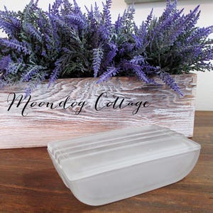 May include: A frosted glass rectangular dish with a raised design on the top. The dish is sitting on a wooden surface. The text "Moondog Cottage" is visible on a wooden box in the background.