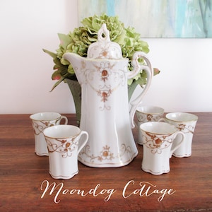 May include: A white porcelain coffee pot with gold floral designs and a matching set of four cups. The coffee pot has a lid with a decorative knob. The cups have a fluted design and are also decorated with gold floral designs.