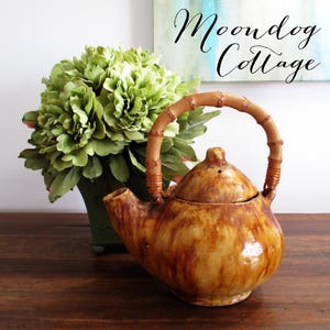 May include: A ceramic teapot with a bamboo handle and a brown, mottled glaze. The teapot is on a wooden surface, with a green floral arrangement in the background. The text "Moondog Cottage" is visible in the background.