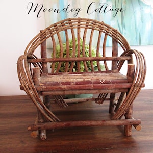 May include: A small, rustic wooden bench made from twigs and branches. The bench has a curved back and armrests, with a seat made of horizontal wooden slats. The text "Moondog Cottage" is written in cursive at the top.