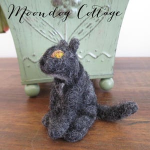 May include: A small, black, felted cat figurine with a yellow eye. The cat is sitting with its tail curled around its body. The figurine is on a wooden surface.