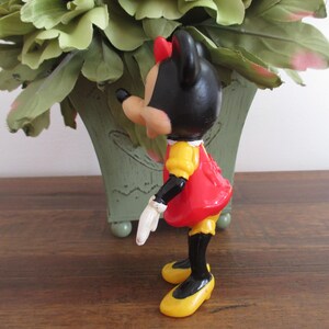 May include: A vintage plastic figurine of Minnie Mouse wearing a red dress with yellow accents and black shoes. She has a black bow on her head and is standing with her right arm outstretched.