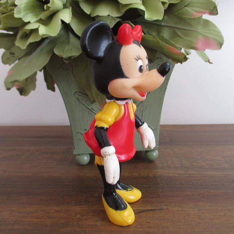 May include: A vintage plastic figurine of Minnie Mouse. She is wearing a red dress with a yellow collar, black shoes with yellow accents, and a red bow.
