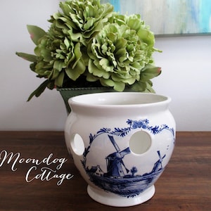 May include: White ceramic bulb pot with blue windmill design. The pot has two large circular openings and a small footed base. The text "Moondog Cottage" is written on the front. Green floral arrangement in the background.