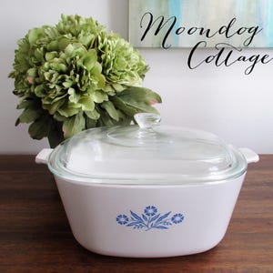 May include: White casserole dish with a clear glass lid. The dish features a blue floral design on the front and has two handles. The text "Moondog Cottage" is written in cursive in the background. A green floral arrangement is in the background.