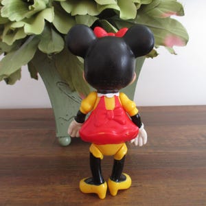 May include: A plastic figurine of Minnie Mouse wearing a red dress with a yellow bodice and a yellow bow. She has black shoes and a black bow in her hair.
