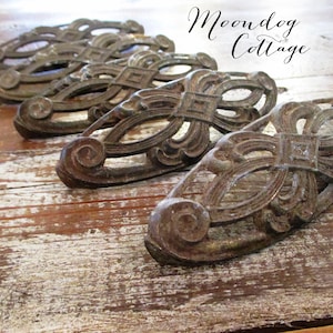 May include: Close-up of several antique bronze drawer pulls with ornate scrollwork and a square center design. The pulls are arranged on a distressed wooden surface, with the text "Moondog Cottage" in the upper right corner.