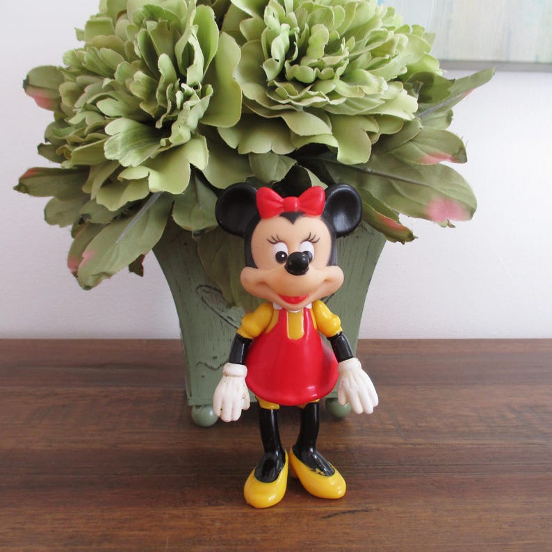 May include: A red and yellow plastic figurine of Minnie Mouse. She is wearing a red dress with a yellow collar and a red bow. She has black shoes and a black nose.