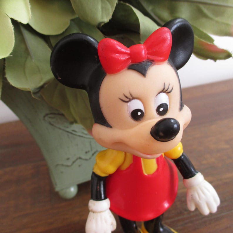 May include: A vintage plastic figurine of Minnie Mouse. She is wearing a red dress with a yellow bodice, a red bow, and white gloves. Her face is painted with black eyes, eyelashes, and a black nose.