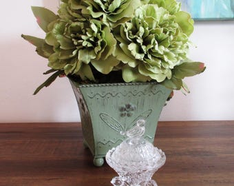 May include: A green metal planter with a distressed finish holds a bouquet of artificial green peonies. A small glass sugar bowl with a bird finial sits on a wooden table in front of the planter.