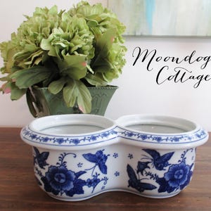May include: A white ceramic double planter featuring a blue floral and butterfly pattern. The oval-shaped vessel has two circular openings and a wide rim. The text Moondog Cottage is visible in the upper right corner above the planter.