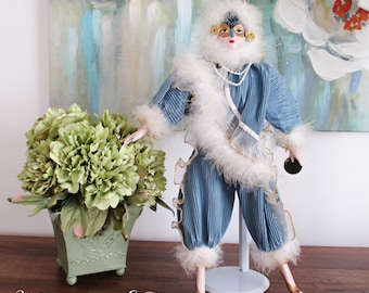 May include: A porcelain doll wearing a blue and white costume with a feathered trim and a masquerade mask. The doll is standing on a white stand and is holding a small black purse. The doll is in front of a painting with abstract brushstrokes.
